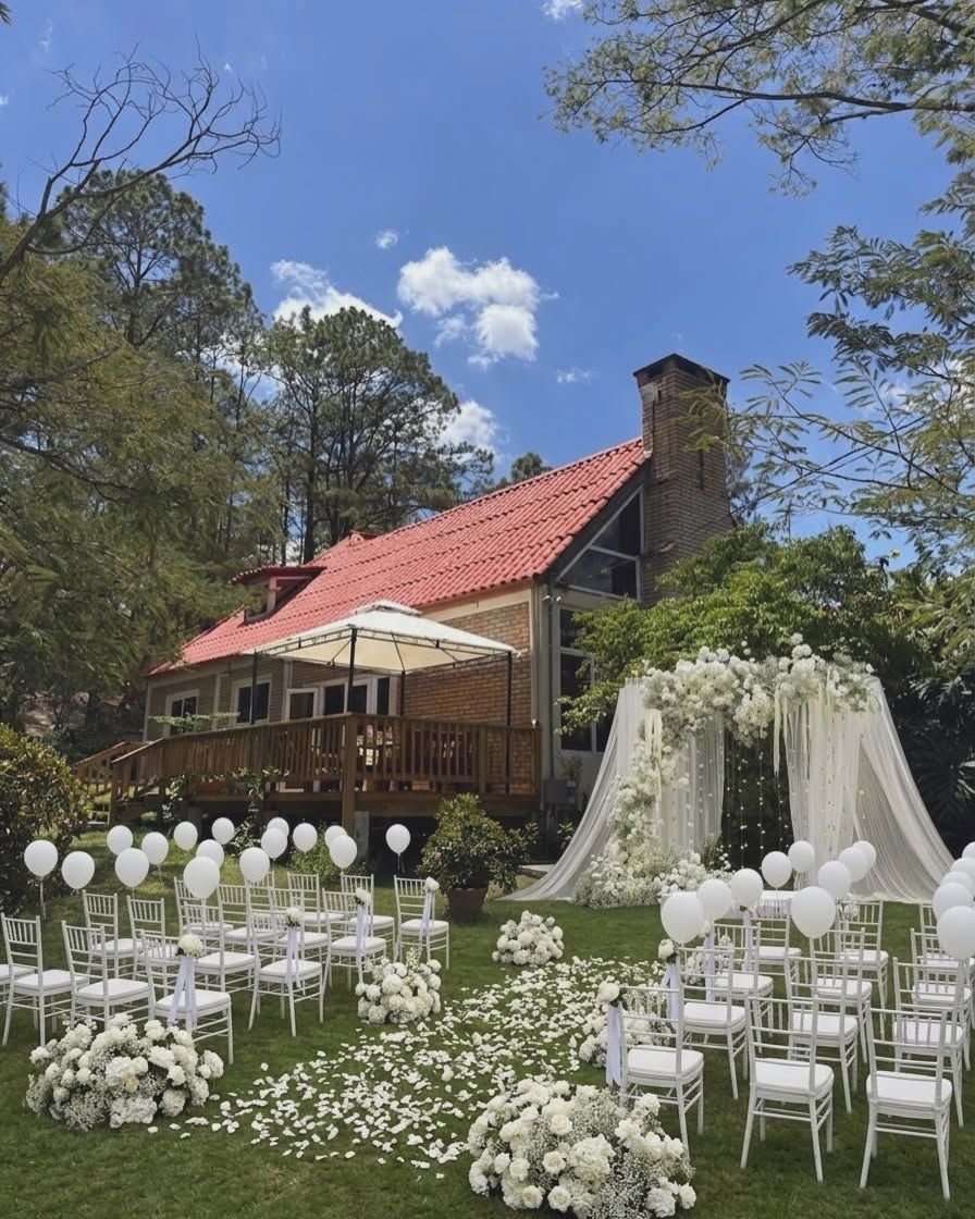 Ceremonia montada en el jardín frente a la casa: sillas blancas, globos y un arco de flores blancas