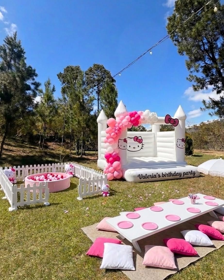 Un brincolín blanco con globos rosados y una mesa baja con cojines en el césped