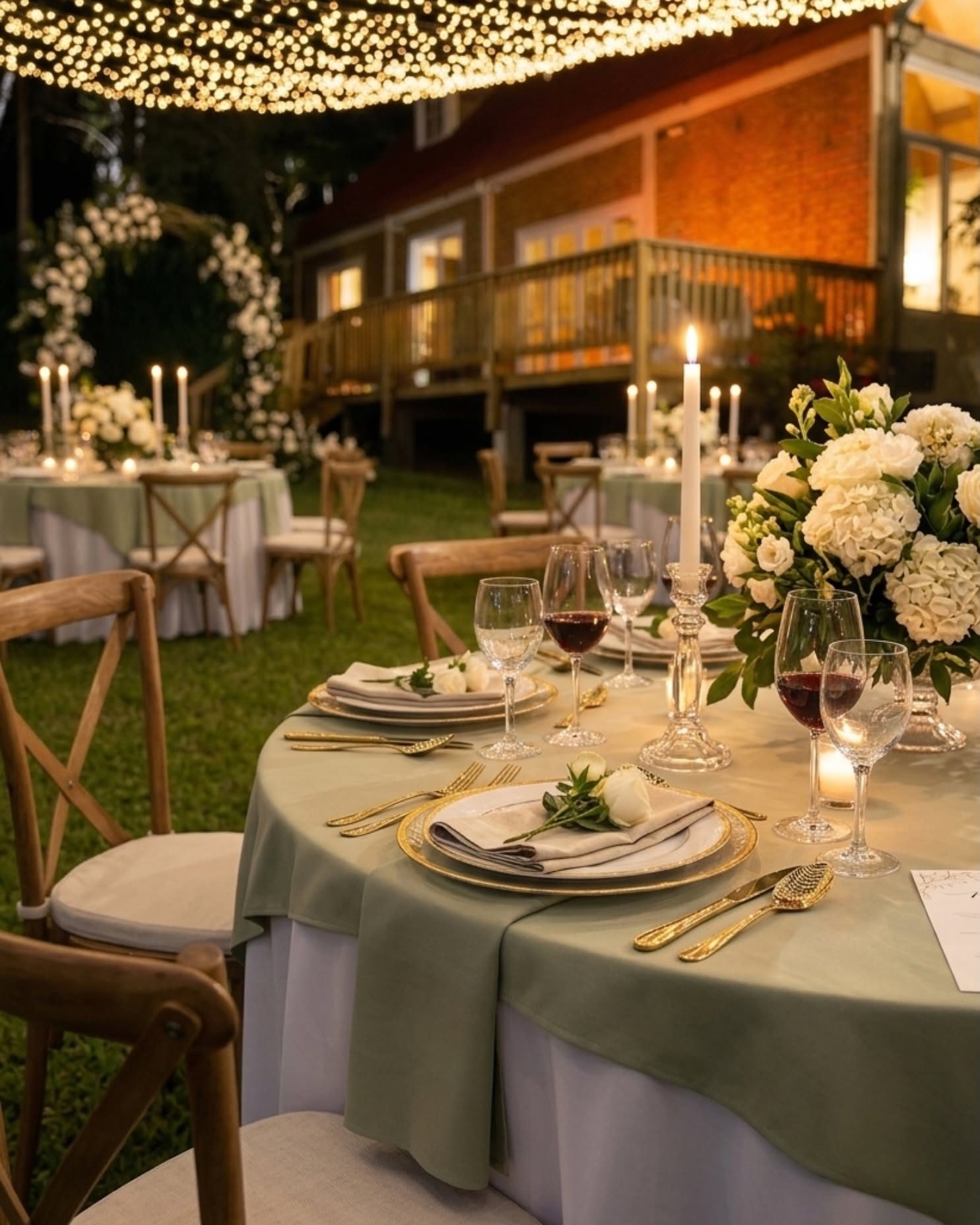 Mesas redondas con velas y flores blancas en el jardín, bajo una red de luces, con la casa al fondo