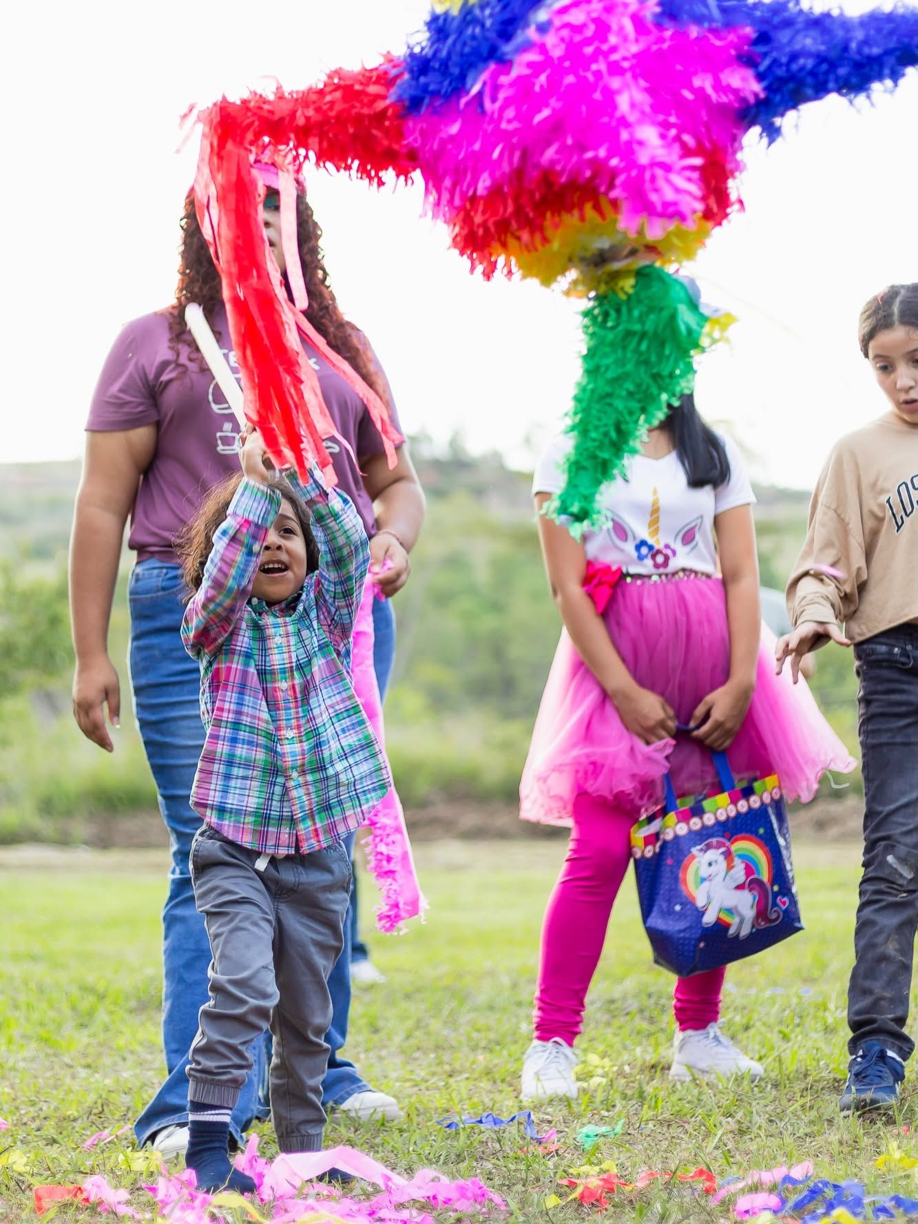 Un niño le pega a la piñata en el césped, rodeado de niñas y adultos