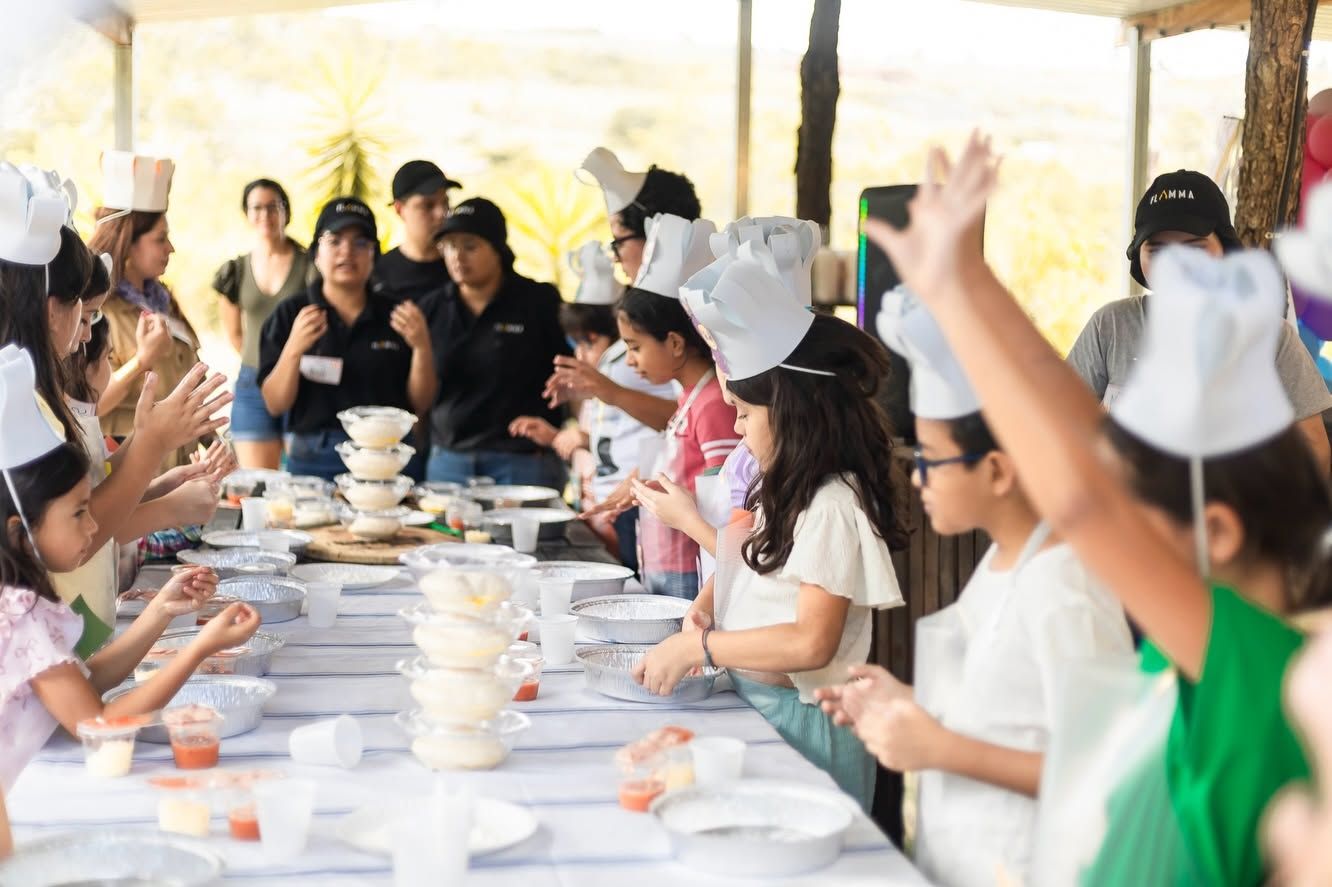 Niños con gorros de chef preparan pizzas en una mesa larga bajo techo